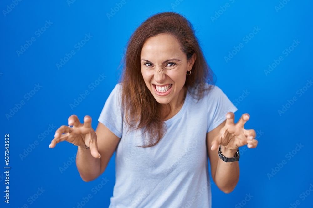 Poster brunette woman standing over blue background smiling funny doing claw gesture as cat, aggressive and