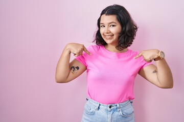 Young hispanic woman standing over pink background looking confident with smile on face, pointing...