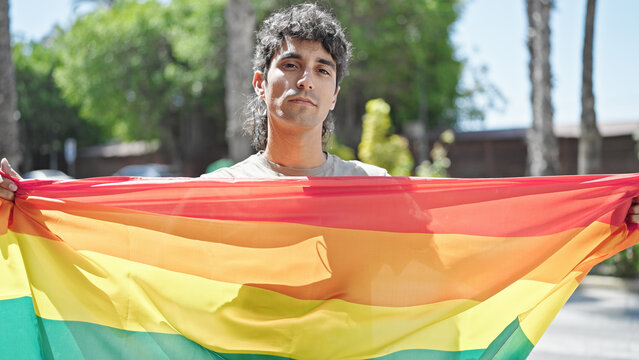 Young hispanic man holding rainbow flag with relaxed expression at street