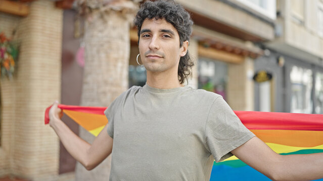Young hispanic man holding rainbow flag with relaxed expression at street
