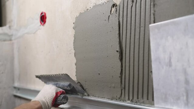 A man puts tiles on the wall. Builder, tiler puts gray ceramic tiles on the wall in the bathroom. Applying adhesive solution on the wall, for gluing tiles in the bathroom, close-up.