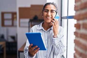 Young beautiful hispanic woman business worker talking on smartphone using touchpad at office