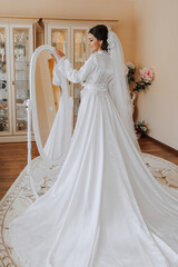 Fototapeta premium Happy dark-haired brunette bride in a satin robe and professional make-up standing in her room near the mirror and getting ready for her wedding day