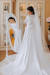 Fototapeta na wymiar Happy dark-haired brunette bride in a satin robe and professional make-up standing in her room near the mirror and getting ready for her wedding day