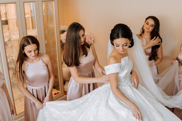 Stylish beautiful bridesmaids in matching silk dresses helping gorgeous brunette bride in white dress get ready for wedding, morning preparations, woman putting on dress - obrazy, fototapety, plakaty