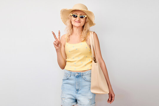 Portrait of positive mature blonde woman in yellow summer clothes showing peace and love gesture with fingers isolated on white studio background