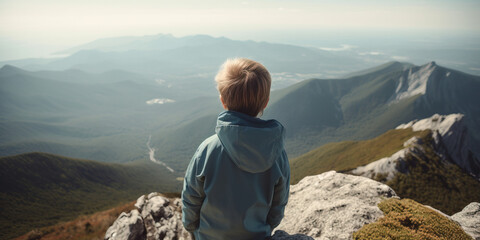 A hiking young blonde fair haired boy with a jacket stares out across a mountain landscape at the top of a high mountain