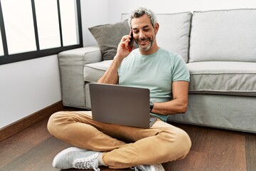 Middle age grey-haired man talking on smartphone using laptop sitting on floor at home