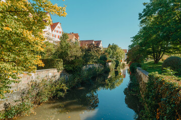 Fototapeta na wymiar Beautiful small river with clean and clear water front of colorful autumn trees and small old town on the hill agaist nice blue and clouds sky during autumn in Europe