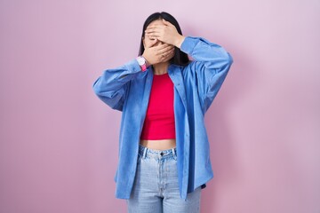 Young asian woman standing over pink background covering eyes and mouth with hands, surprised and shocked. hiding emotion