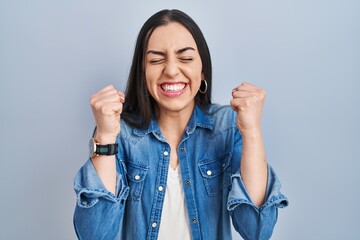 Hispanic woman standing over blue background excited for success with arms raised and eyes closed celebrating victory smiling. winner concept.