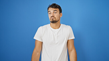 Young hispanic man standing clueless over isolated blue background