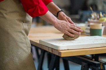 Unrecognizable woman wedging clay with hands standing behind table in studio.