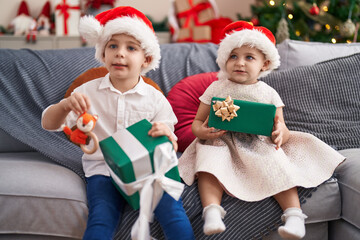 Two kids holding gifts sitting on sofa by christmas tree at home