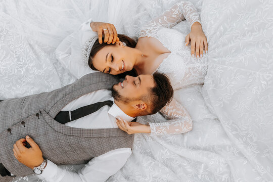 Fashionable groom and cute brunette bride in white dress with crown, hugging, laughing in park, garden, outdoors. Wedding photography, portrait of smiling newlyweds.
