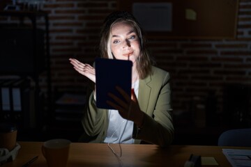 Blonde caucasian woman working at the office at night pointing aside with hands open palms showing copy space, presenting advertisement smiling excited happy