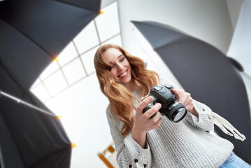 Portrait of happy redhead girl smiling and holding professional digital camera, Captivating image of a young professional woman posing with camera gear in a well-equipped studio illuminated by lights.