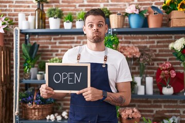 Young hispanic man working at florist holding open sign puffing cheeks with funny face. mouth...
