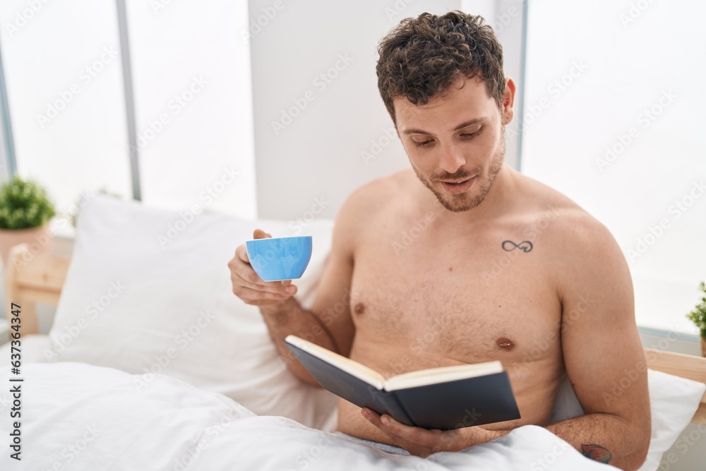 Wall mural Young hispanic man reading book and drinking coffee sitting on bed at bedroom