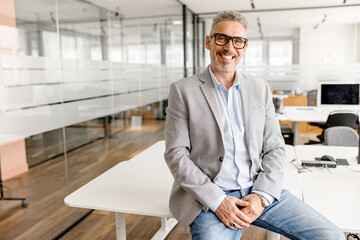 Successful good-looking modern mature businessman leaned on the desk in contemporary office, middle-aged male employee, small business owner, investor in suit looking at the camera, proud entrepreneur