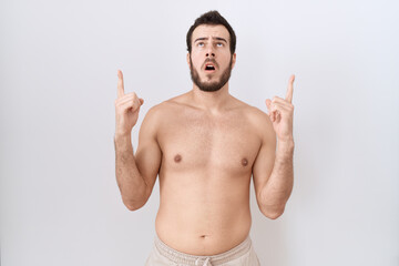 Fototapeta premium Young hispanic man standing shirtless over white background amazed and surprised looking up and pointing with fingers and raised arms.