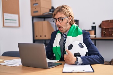 Caucasian man with mustache working at the office supporting football team scared and amazed with...
