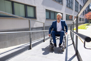 Gray haired businessman in formal wear gets to work in wheelchair rides ramp.