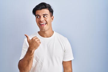 Hispanic man standing over blue background smiling with happy face looking and pointing to the side with thumb up.