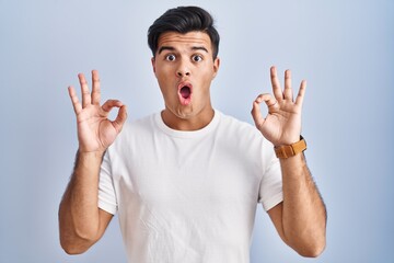 Hispanic man standing over blue background looking surprised and shocked doing ok approval symbol...