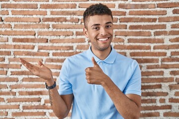 Brazilian young man standing over brick wall showing palm hand and doing ok gesture with thumbs up,...