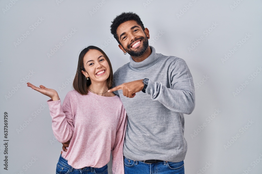 Poster Young hispanic couple standing together amazed and smiling to the camera while presenting with hand and pointing with finger.