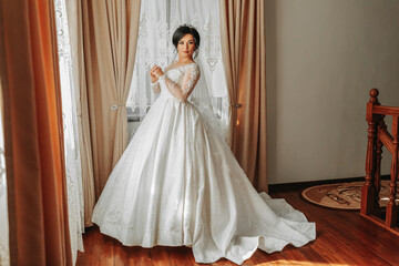 Wedding. Bride in a beautiful dress standing by the window indoors in an expensive interior at home. Trendy wedding style full length shot. Young attractive Caucasian brunette model as a bride