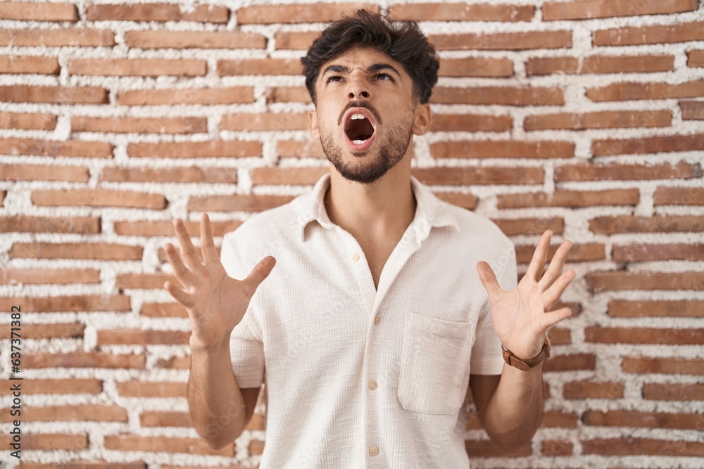 Poster arab man with beard standing over bricks wall background crazy and mad shouting and yelling with agg