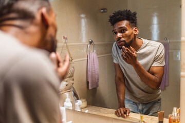 Homem jovem de cabelo jovem conferindo sua barba no espelho do banheiro.