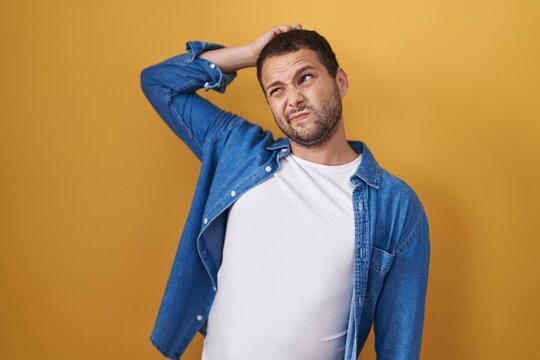 Hispanic man standing over yellow background confuse and wondering about question. uncertain with doubt, thinking with hand on head. pensive concept.
