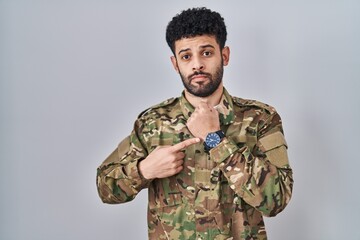 Arab man wearing camouflage army uniform in hurry pointing to watch time, impatience, looking at the camera with relaxed expression