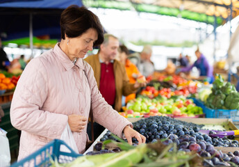 Smiling elderly female customer choosing organic plums while walking at local bazaar