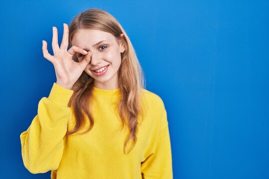 Young caucasian woman standing over blue background doing ok gesture with hand smiling, eye looking through fingers with happy face.