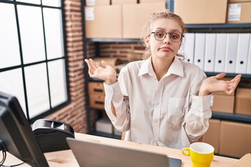 Young caucasian woman working at small business ecommerce using laptop clueless and confused expression with arms and hands raised. doubt concept.