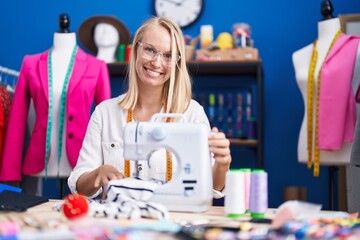 Young blonde woman tailor smiling confident using sewing machine at sewing studio