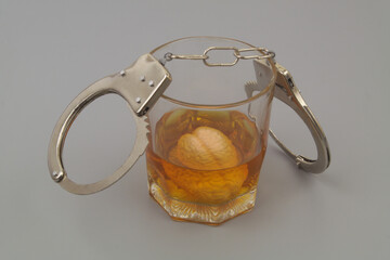 Alcoholism and crime concept. Glass of alcohol with human brain and handcuffs on gray background.