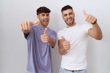 Homosexual gay couple standing over white background approving doing positive gesture with hand, thumbs up smiling and happy for success. winner gesture.