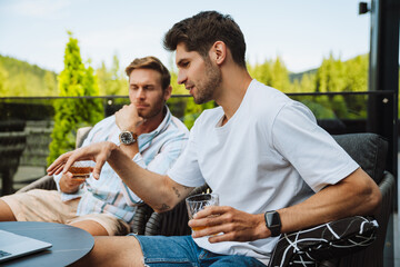 Two male friends drinking whiskey and using laptop while relaxing on terrace in mountains
