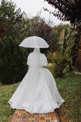 Beautiful stylish bride with a white umbrella, walks in the rain in the park, wedding photo session in the rain. A model with an umbrella in a lush wedding dress