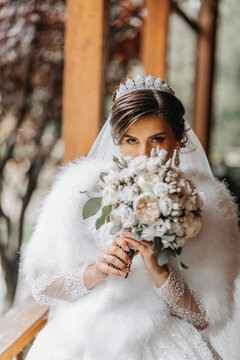 A beautiful queen with a bouquet of flowers in her hands. Charming woman bride is walking in the park. A wedding outfit, a lush dress, a luxurious cloak, a white cape made of swan feathers.