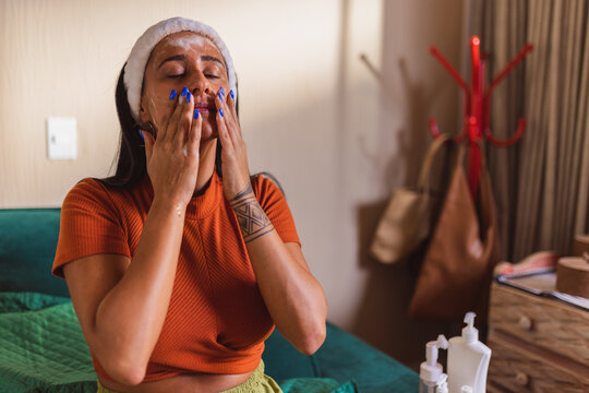 Jovem mulher indigena, sentada na cama, aplicando hidratante no rosto.