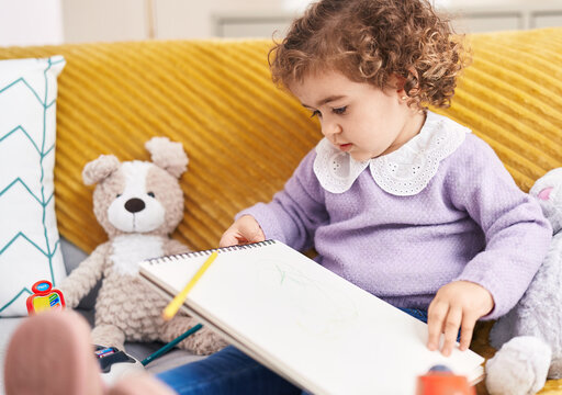 Adorable hispanic girl drawing on notebook sitting on sofa at home