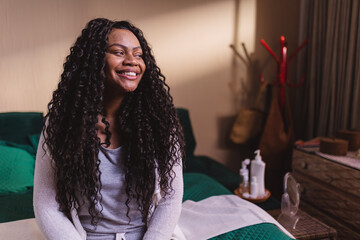Linda mulher de cabelos cacheados, sorrindo, sentada na cama vestindo pijama.
