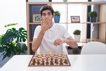 Young hispanic man playing chess sitting on the table covering mouth with hand, shocked and afraid for mistake. surprised expression