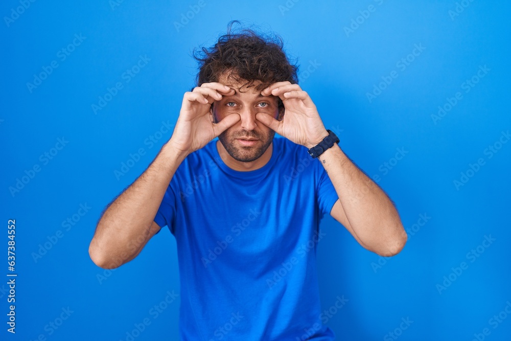 Canvas Prints Hispanic young man standing over blue background trying to open eyes with fingers, sleepy and tired for morning fatigue